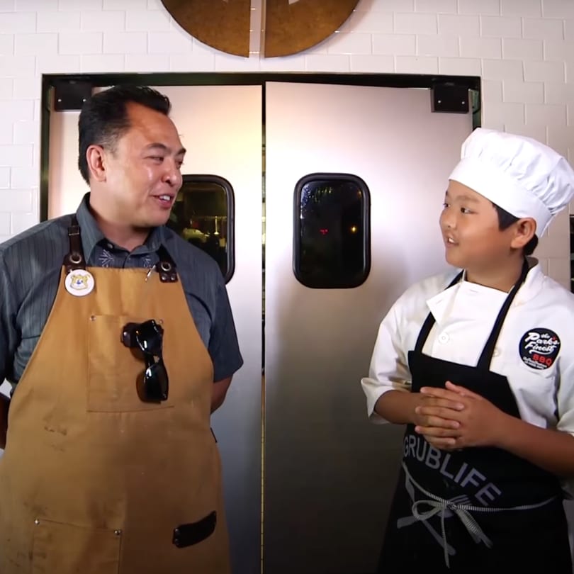 An adult man in an apron talks to a young boy in a chef's hat and apron.