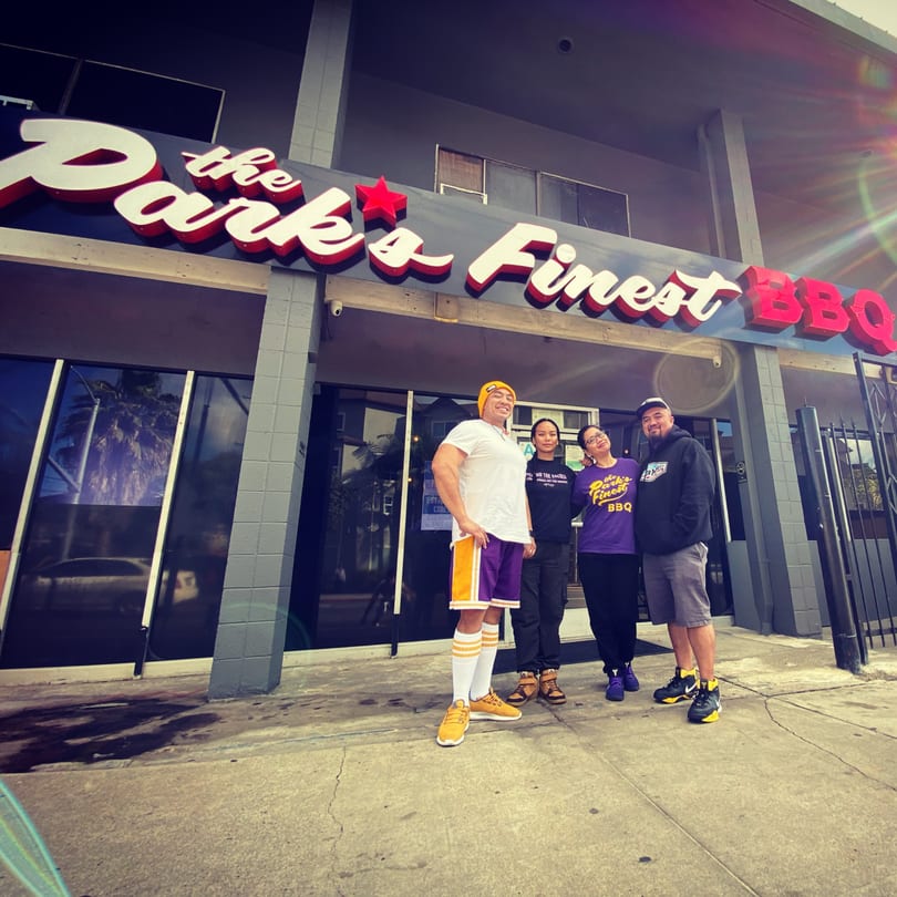 Four people stand smiling in front of The Park's Finest BBQ restaurant, with a bright sun flare.