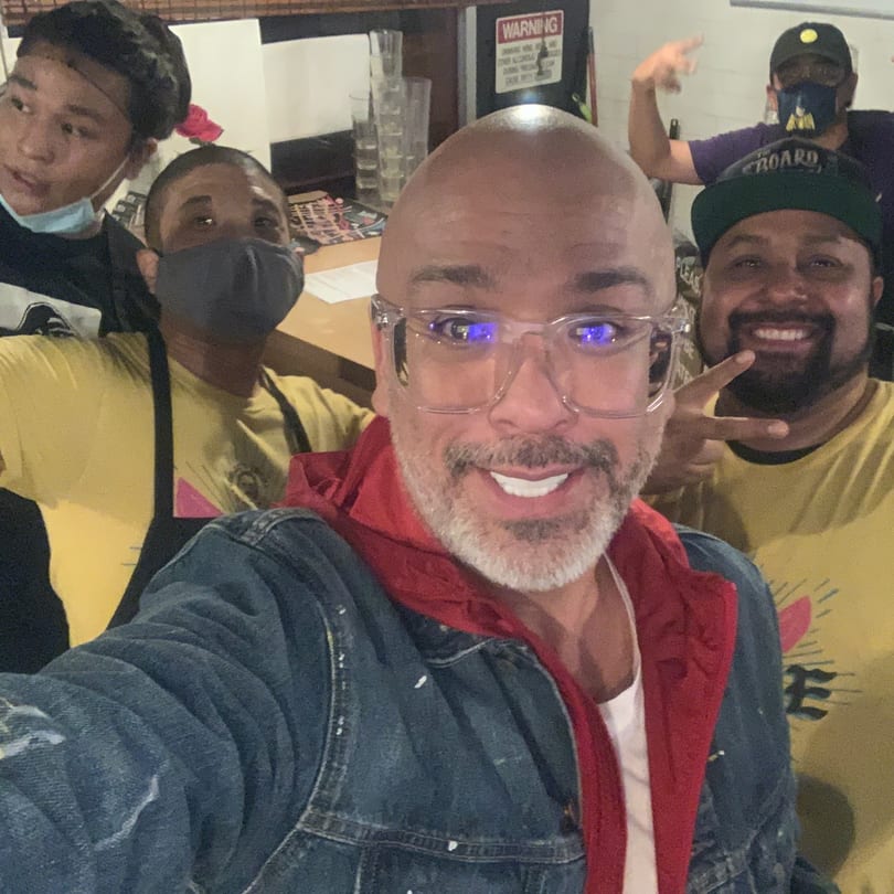 A man in clear glasses and a red hoodie takes a selfie with four smiling staff members.
