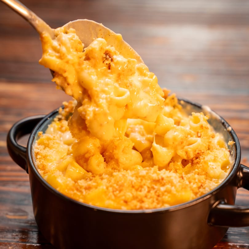 Spoon lifting creamy mac and cheese with a crispy topping from a black bowl on a wooden table.