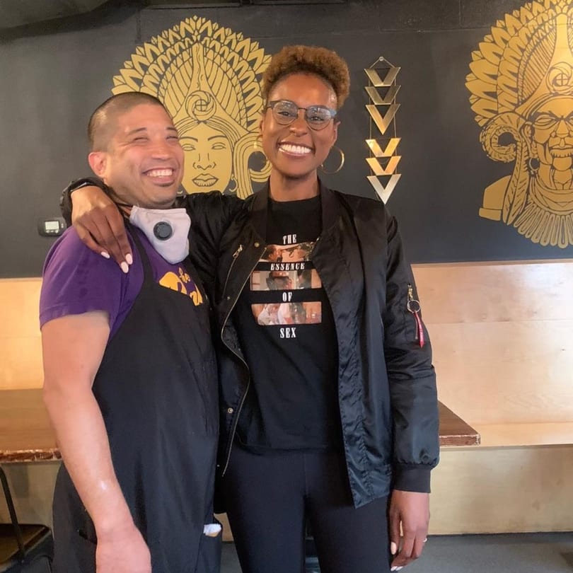 Two smiling people, one in a purple shirt and apron, the other in a black jacket, embrace.