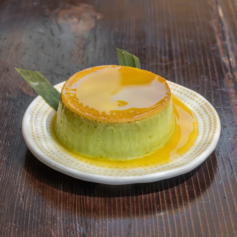 A green flan dessert with yellow caramel sauce on a white plate with green leaf garnish.