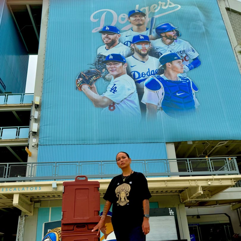 A woman stands in front of a large Dodgers mural at a stadium, with red food containers beside her.