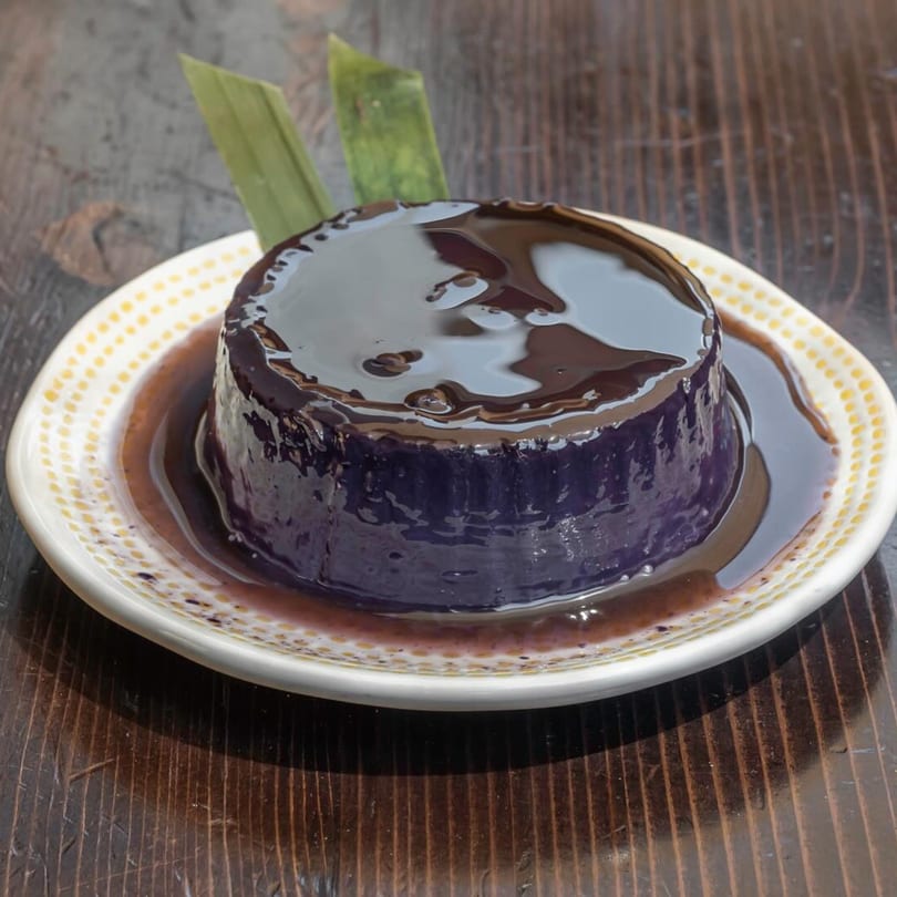 Purple ube panna cotta with dark chocolate sauce and green garnish on a white plate.