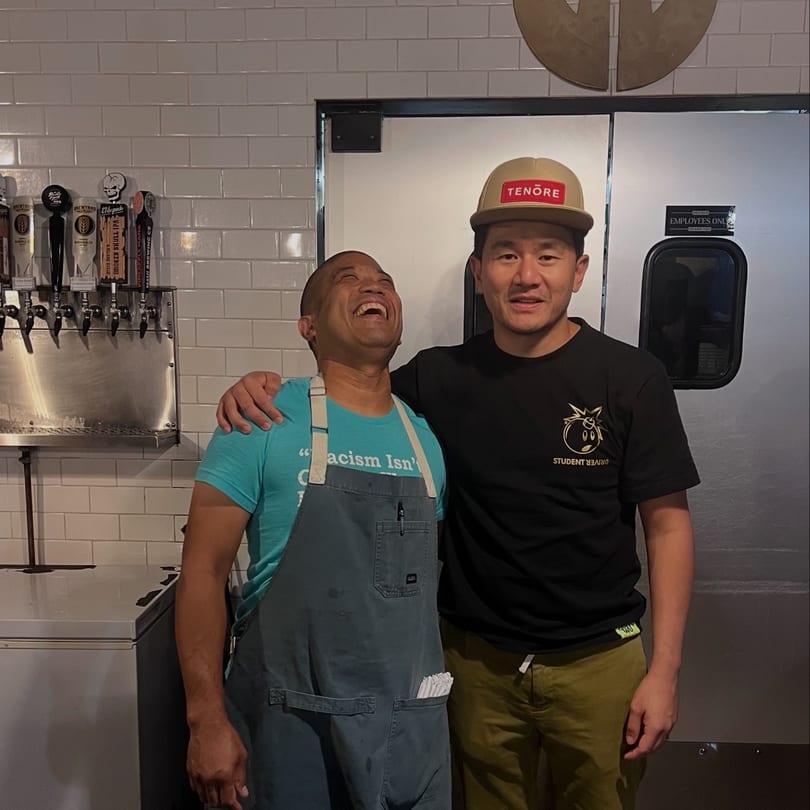 Two men, one laughing, stand in front of beer taps and a white tiled wall at The Park's Finest.