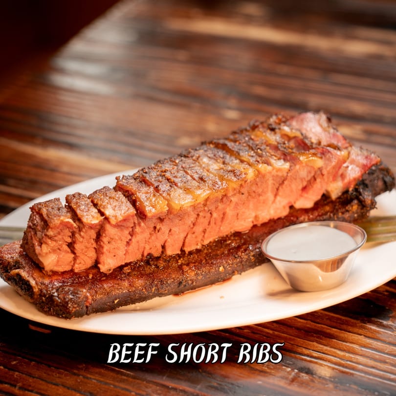 Sliced beef short ribs with a side of white sauce on a white plate, on a wooden table.